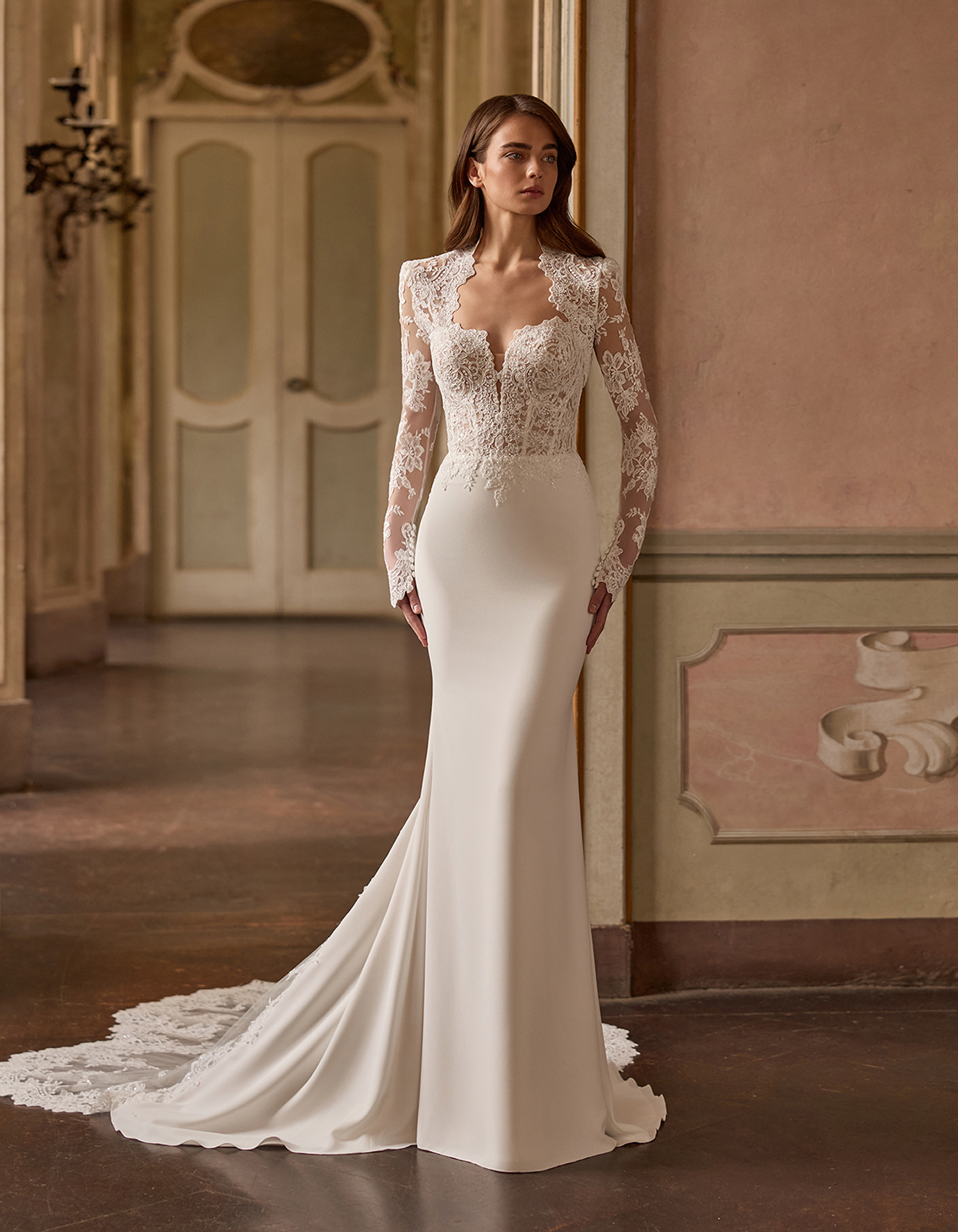 Bride wearing a white lace wedding gown with long sheer sleeves and a flowing train in an ornate hallway.