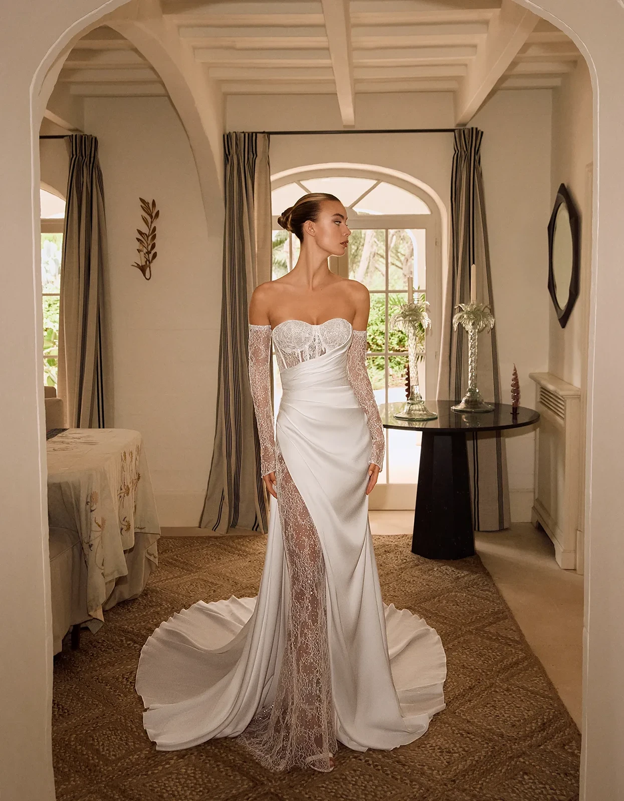 Bride in a white off-the-shoulder lace wedding gown with long lace sleeves, standing in a bright, elegant room with a sweeping train on the floor.