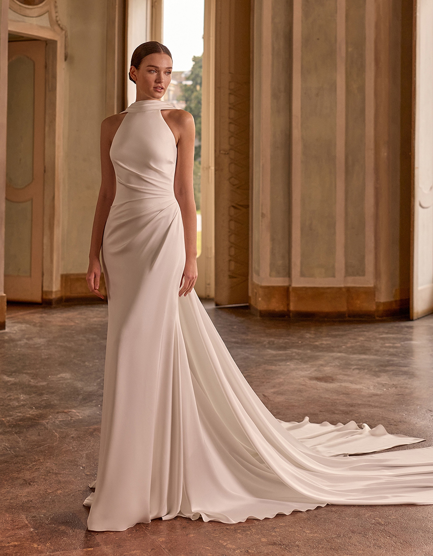 Woman in a white, high-neckline wedding gown with a long train standing in a historic marble hallway.