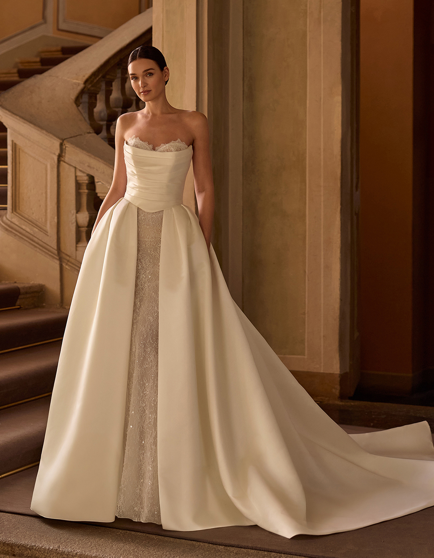 Bride wearing a strapless white wedding gown with a lace bodice, standing on a grand staircase in a warm, historic interior.