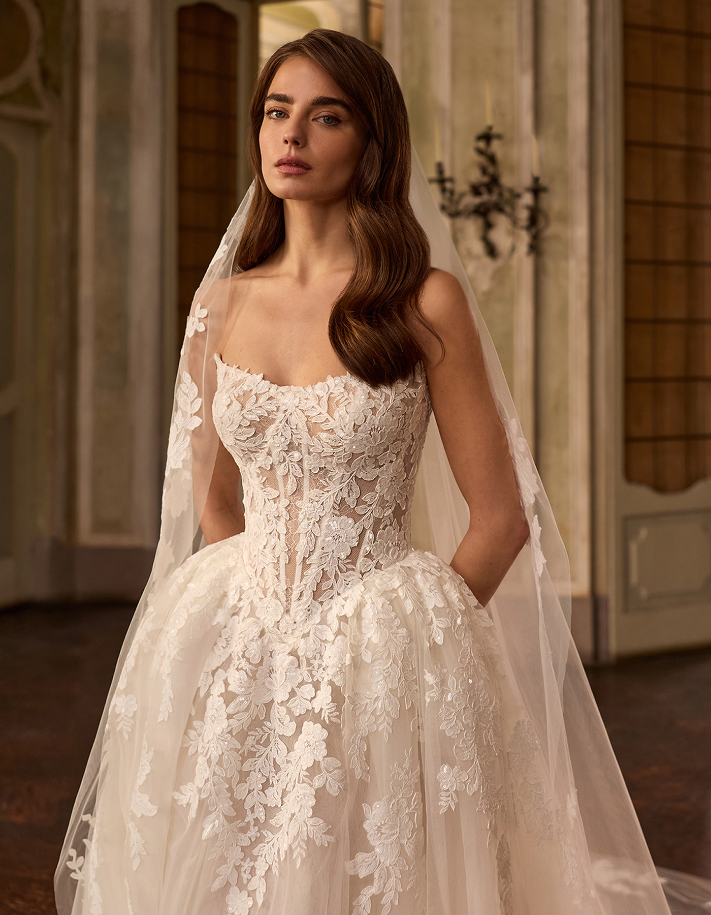 Bride wearing a lace wedding gown with a long veil, posing indoors in a grand hall.