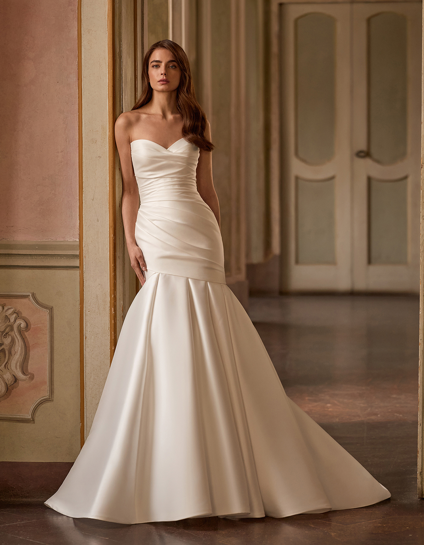 Woman in a strapless white wedding dress standing against a vintage wall in an ornate hallway.
