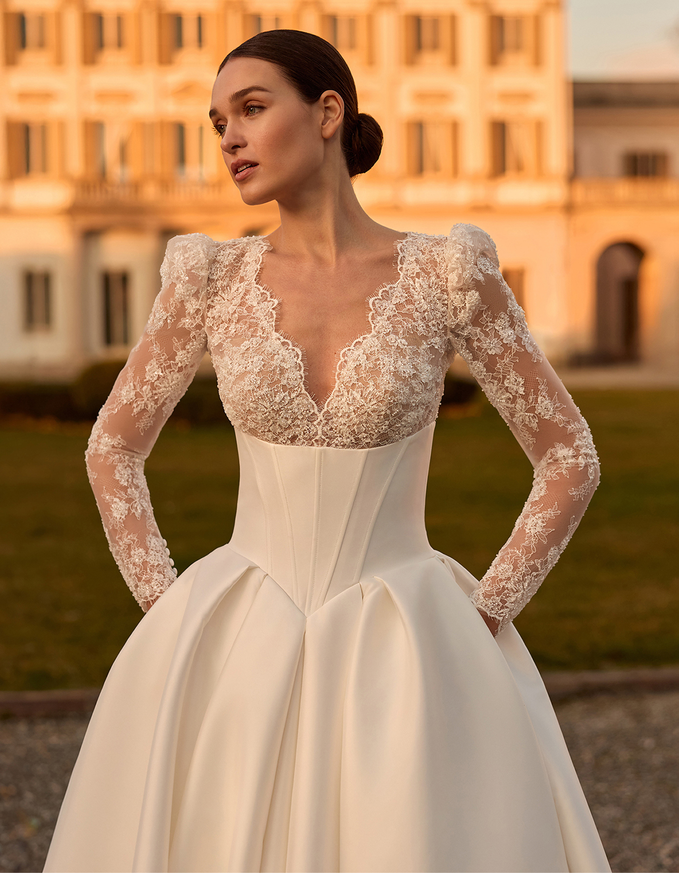 Bride in a lace long-sleeve wedding gown posing outdoors at sunset with a grand building in the background